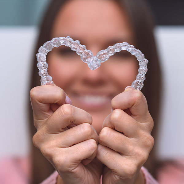 The image shows a person holding up two hands, each displaying a clear, heart-shaped object that resembles a dental retainer or aligner. The individual is smiling and has their hair pulled back, revealing the face partially. They are wearing a light pink top and are standing against a neutral background with a blurred white element on the right side.