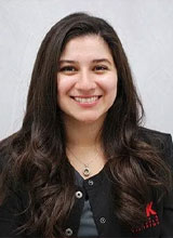 The image shows a smiling woman with long hair, wearing a dark top and a name tag. She is posing for a portrait against a neutral background.
