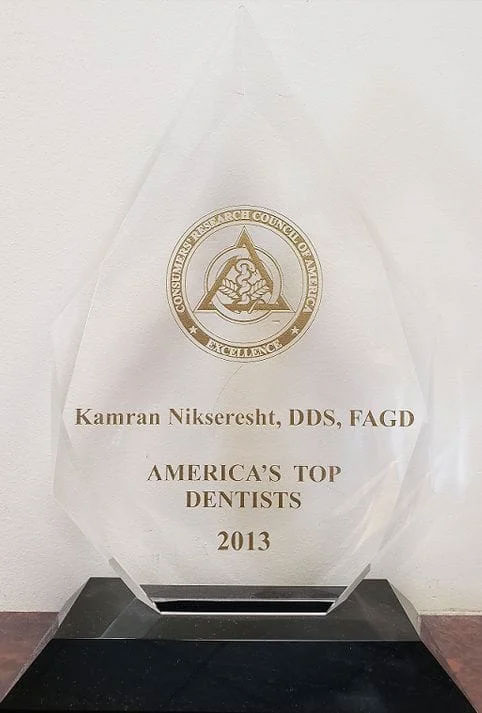The image shows a plaque with text, awarded to an individual named Kamran Nikseresht, recognizing their achievement as America's Top Dentists 2013. The plaque is placed on a table and features a logo, possibly representing the awarding organization.
