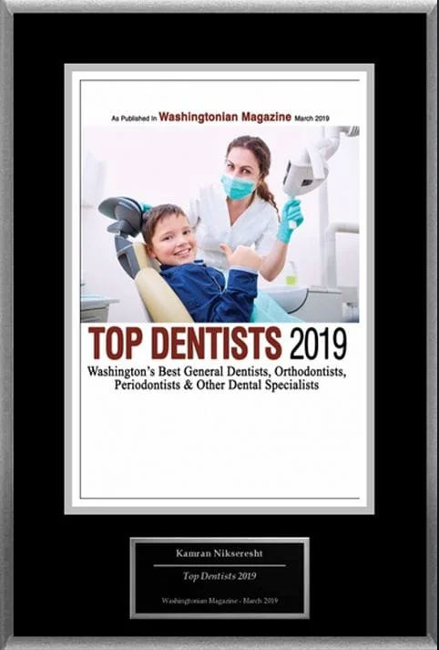 This is a framed certificate or award, possibly for an achievement in dentistry or dental education, featuring a photograph of a woman wearing protective eyewear and a surgical mask while sitting at a dental chair, with a young child seated on her lap. The image includes the text  TOP DENTISTS 2019  and mentions  WASHINGTON S BEST GENERAL DENTISTS   ORTHODONTISTS   PERIODONTISTS.  There is also an emblem at the bottom left corner that says  KAMIN NIKOWSKI TOP DENTIST MARCH 2019,  indicating the recipient of the award. The certificate has a black border and is set against a white background.