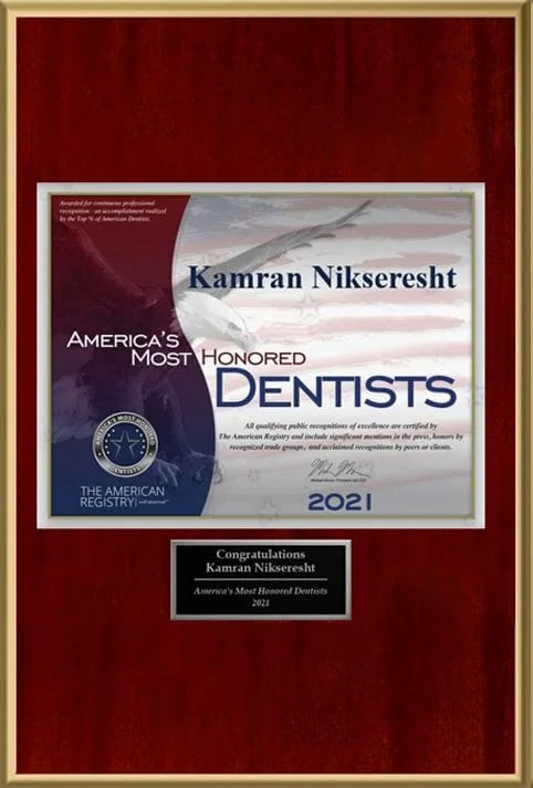 The image shows a framed certificate with text that reads  KAMRAN NIKSERST  and  AMERICA S MOST HONORED DENTISTS 2021,  alongside an American flag, a bird graphic, and a red seal. Below the main text is a smaller inscription stating  THE AMERICAN COLLEGE OF DENTAL RADIOLOGY.  The certificate is displayed on a wooden surface with a plaque that reads  CONGRATULATIONS KAMRAN NIKSERST  at the bottom.